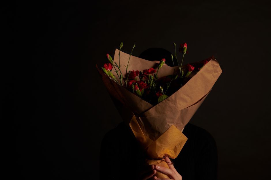 A person holding a bouquet of fresh red and pink carnations wrapped in brown craft paper, with the flowers and stems visible and vibrant. The bouquet is held close to their chest, partially obscuring 