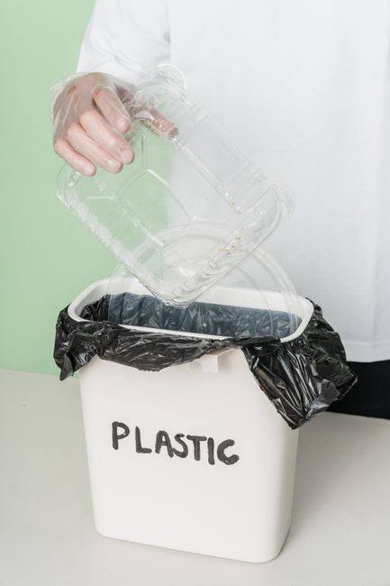A person wearing a white long-sleeved shirt is seen discarding a clear glass flower vase into a white recycling bin lined with black plastic, labeled 'PLASTIC'. The scene is set against a light green 