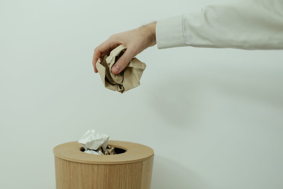 A person wearing a white long-sleeve shirt is disposing of crumpled brown and white paper into a wooden trash bin, with an opening at the top. The background is plain and light-colored, and the scene 