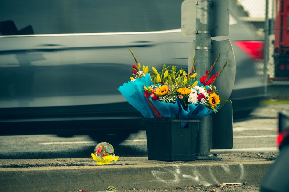 A vibrant bouquet of fresh flowers arranged in a black plastic container, placed on the sidewalk near a street pole. The arrangement features yellow sunflowers, white daisies, red gladiolus, and green