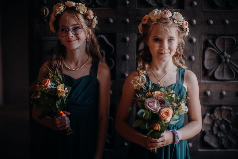 Two young girls wearing teal dress and floral crowns made of pale pink and cream roses, along with other mixed blooms and greenery, are holding floral bouquets. The girl on the left, with glasses and 
