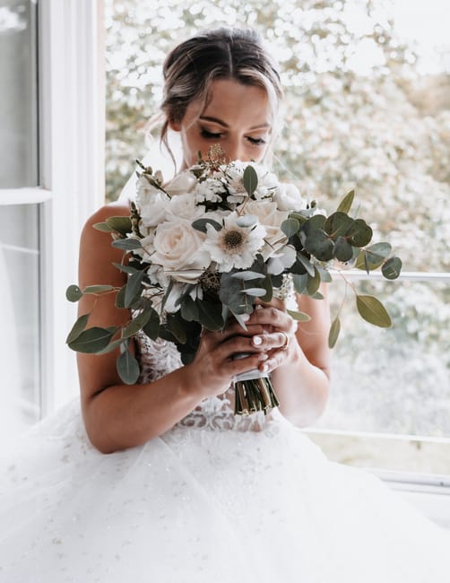 A bride in a white wedding dress with lace detailing holds a large floral bouquet close to her face, gently smelling the fresh flowers. The bouquet includes white roses, delicate daisies, and eucalypt