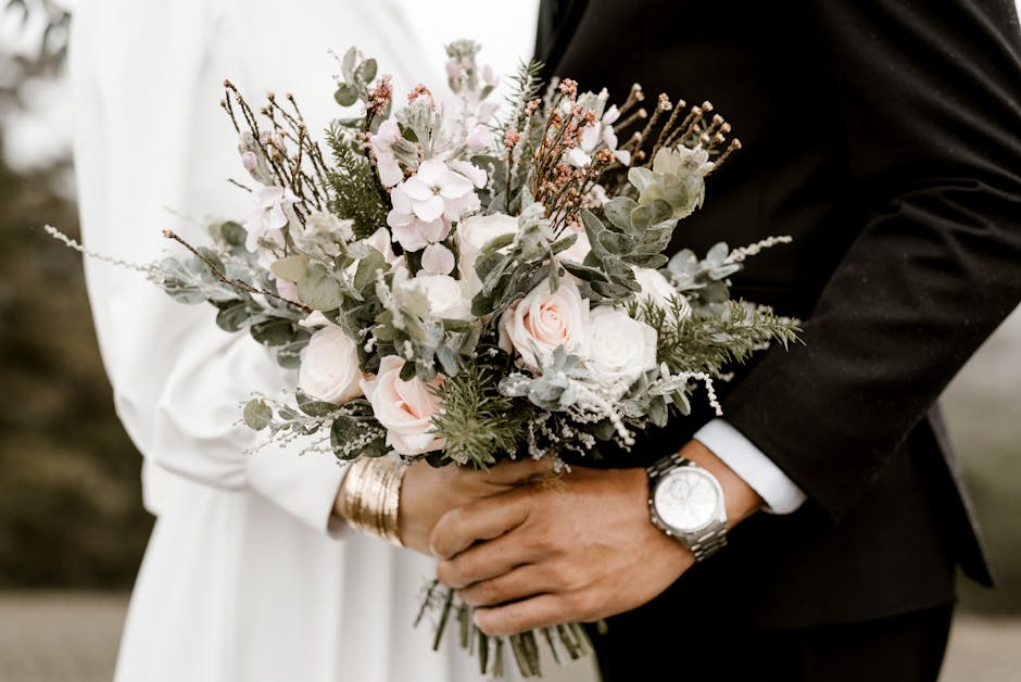 A close-up of a bride and groom holding a romantic floral bouquet together. The bouquet features soft pink roses, white hydrangeas, and sprigs of eucalyptus and other greenery, arranged in a loose, na