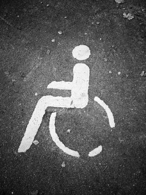 A close-up image of a white painted wheelchair accessibility symbol on a dark asphalt surface, featuring a simplified figure sitting in a wheelchair with circular wheels and an outline of the person. 