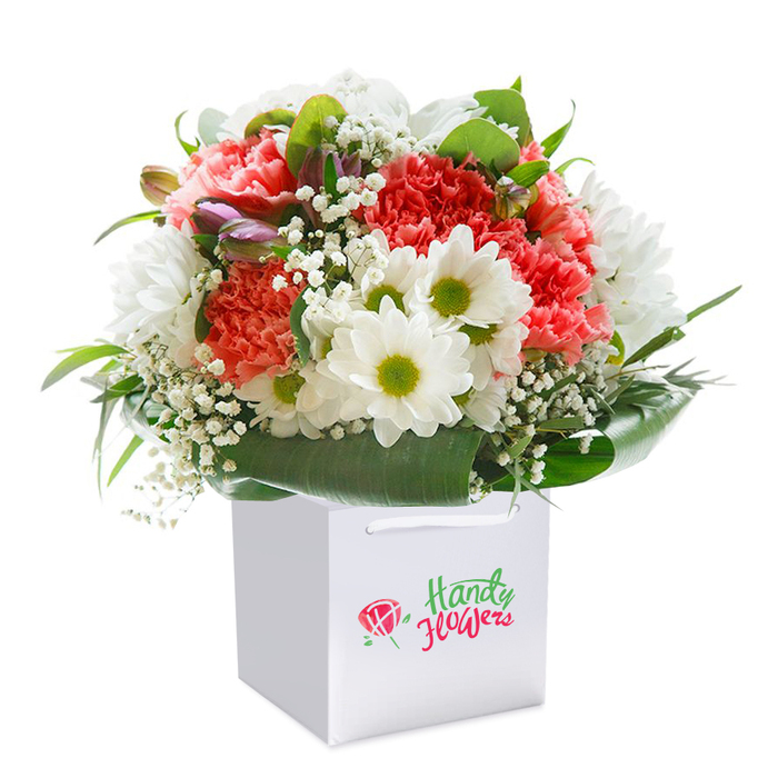 Set against a seamless white canvas, this vibrant bouquet is expertly arranged in a sleek, square white gift bag, embodying both elegance and sentimentality-true to the spirit of South Harrow's craft floristry. The eye is immediately drawn to the plush, coral pink and peach carnations, every petal ruffled and lush, exuding a sense of comforting abundance. Tucked gracefully among them, crisp white daisies with bright yellow-green centres burst forth, injecting cheer and freshness much like the morning sun rising over Harrow-on-the-Hill. Threads of delicate baby's breath weave through the blooms, their cloud-like clusters bringing a gentle softness and refined detail to the arrangement. Along the left, a few unopened buds-brushed with hints of purple and pink-add both dimension and anticipation, suggesting a moment of beauty yet to unfold. The ensemble is embraced by a halo of rich, deep green foliage, each leaf broad and glossy, forming an elegant natural border around the floral medley. Completing the scene, sturdy white ribbon handles rise from the bag's rim, promising convenient delivery across South Harrow and lending a subtle note of luxury. The composition's full, rounded silhouette and luminous hues encapsulate the freshness and care one expects from a reputable neighbourhood florist. This image embodies a gift made to mark milestones-anniversaries, milestone birthdays, or spontaneous celebrations-capturing both the artistry and heartfelt generosity that define flower giving in South Harrow's close-knit community.